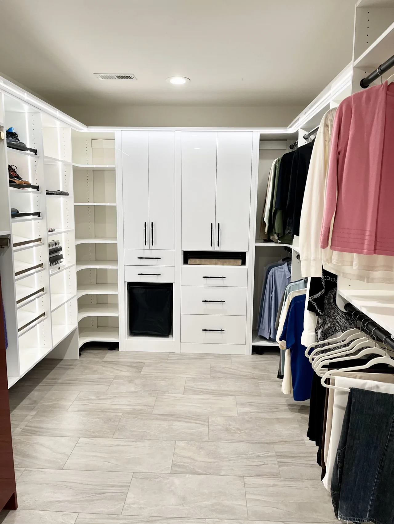 White walk-in closet with lighted shoe shelves, rounded corner shelving, a tall door-and-drawer unit with a built-in hamper, and double hanging