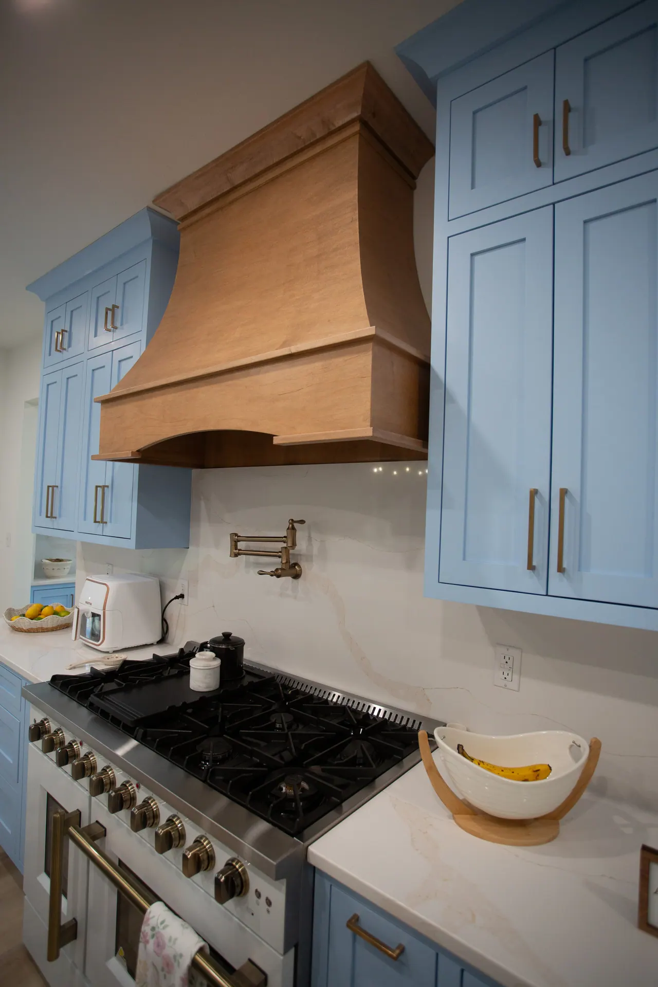 Natural wood range hood and pot filler flanked by blue upper cabinets with brass pulls