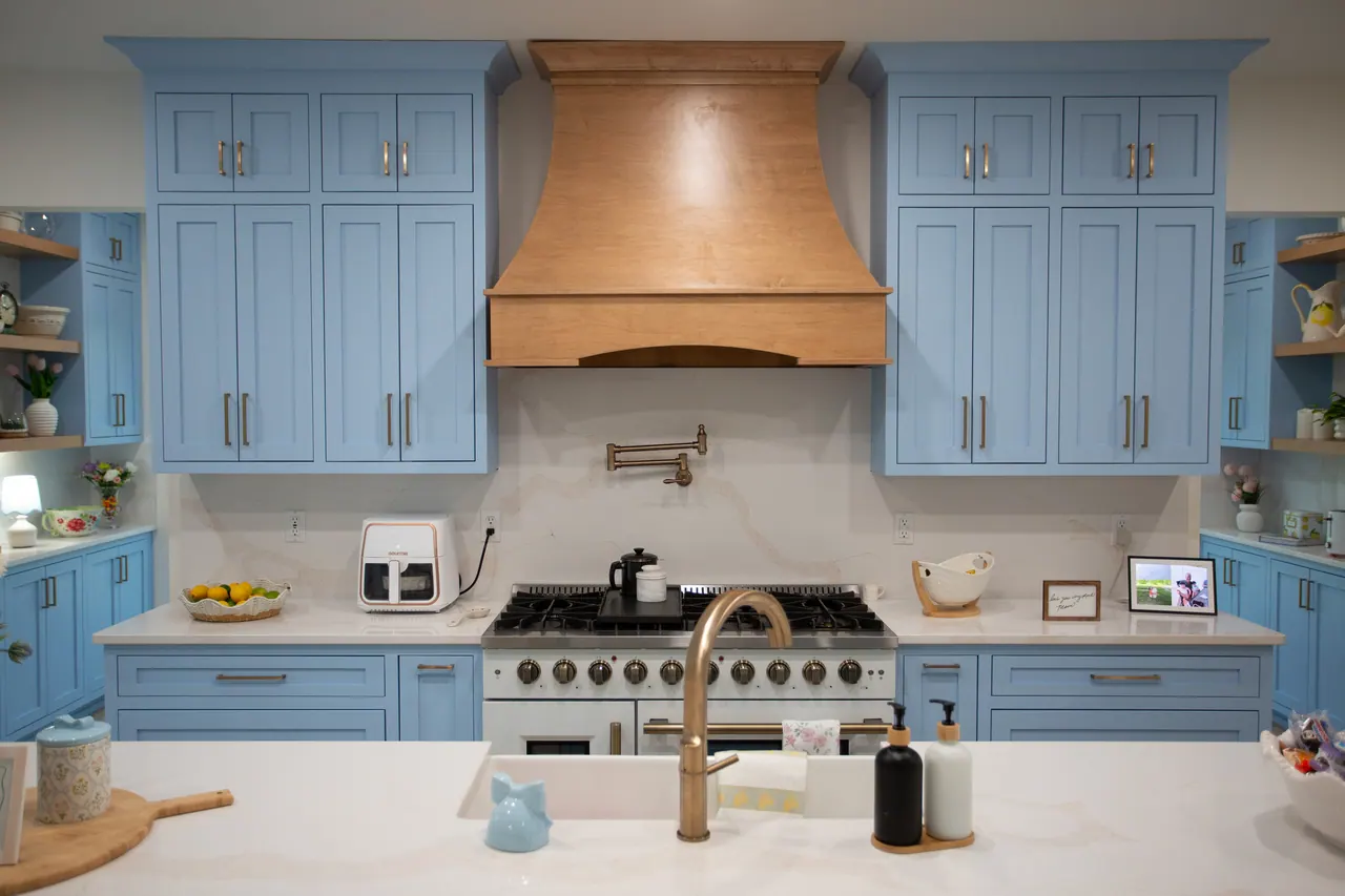Soft blue kitchen cabinets framing a natural wood range hood with gold faucet and white quartz island